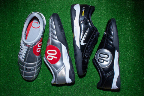 Two pairs of Nike Total 90 III lay on the green floor. One's color is metallic silver, the other is classic black