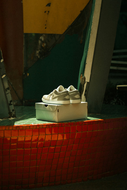 Pair of grey and white Air Jordan 1 Low sneakers displayed on top of a shoebox, placed on a tiled red and green surface in a moody, urban setting with dramatic lighting